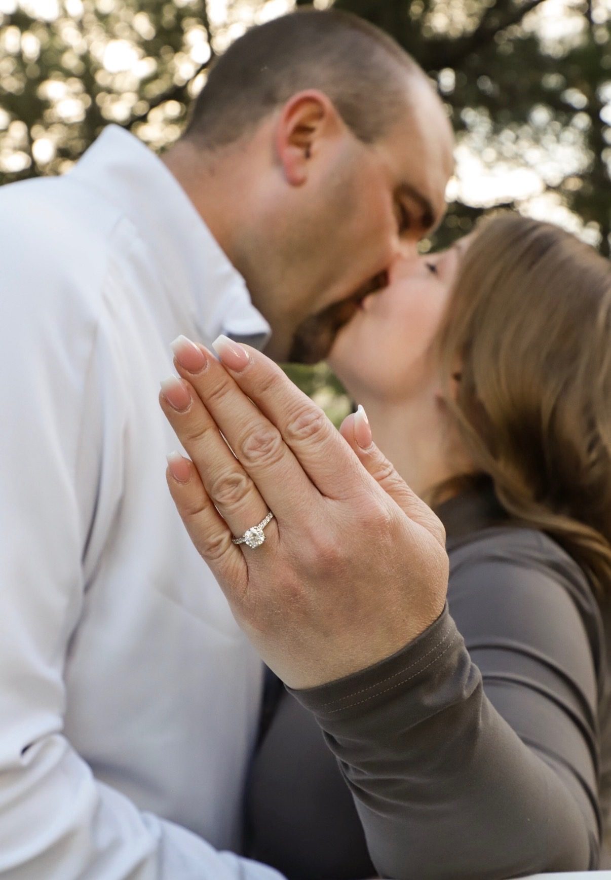 A close-up of a woman's hand showing her engagement ring while she and her fiance kiss behind it.