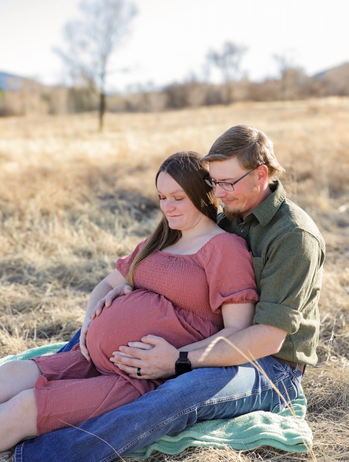 An expectant couple sitting together on a blanket in a dry winter field, both hands resting on her bump.